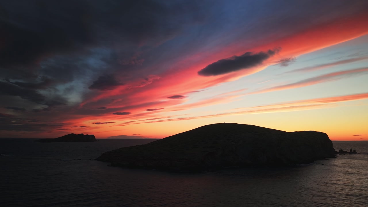 Drone panning around Illa des Bosc in Ibiza at sunset with dramatic colorful sky and ocean.