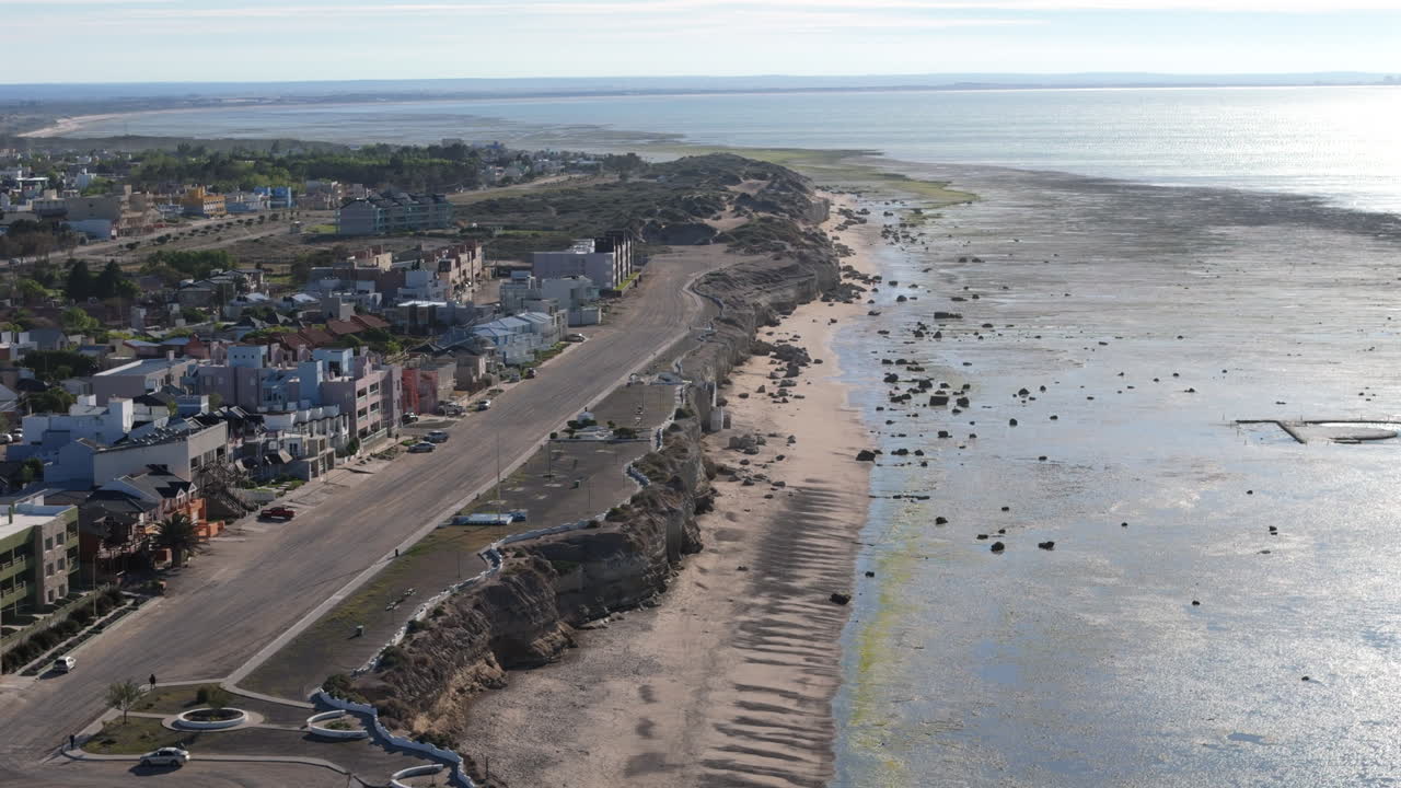 A serene aerial view of Las Grutas, featuring the residential coastline. The image highlights a road stretching along the beach, with rocky formations dotting the shoreline.