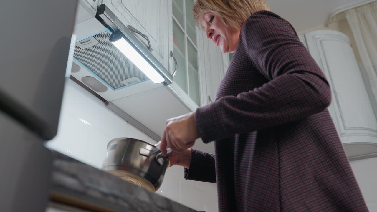 즐거운 여자가 요리를 할 때 춤을 추고, 화려한 옷을 입고, 현대적인 부에서 비를 들고, 캐비 아래의 따뜻한 조명은 그녀가 식사를 준비하는 것을 즐기면서 행복한 표정을 밝히고 있습니다.