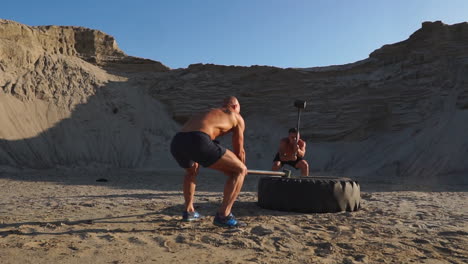 two male athletes training together hit the wheel with a hammer at sunset in the mountains on the sand. endurance training