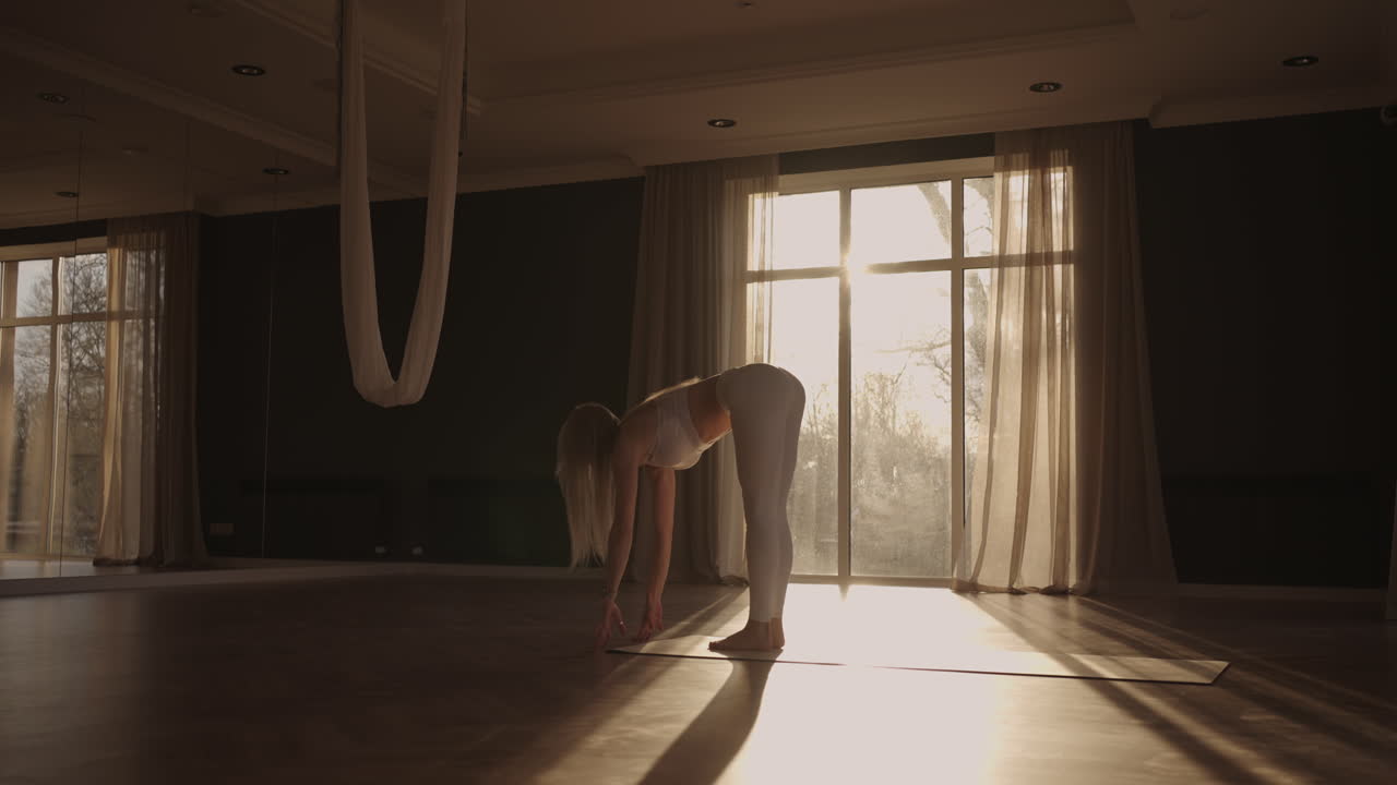 En ung kvinde i hvidt sportstøj strækker ud i en stor hal med store vinduer, filmet i slow-motion, mens solens stråler skinner ind gennem vinduet. Sund livsstil, sund morgen