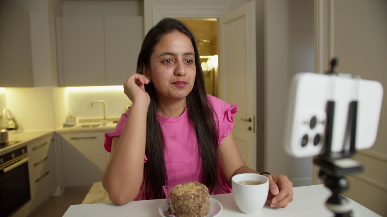 una feliz chica morena en un vestido rosa se sienta en una mesa delante de ella con un pequeño pastel y una taza de café blanco. la chica se comunica a través de video con sus espectadores y amigos en un apartamento moderno en casa