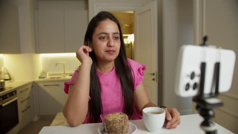 A-happy-brunette-girl-in-a-pink-dress-sits-at-a-table-in-front-of-her-with-a-small-cake-and-a-mug-of-white-coffee.-The-girl-communicates-via-video-with-her-viewers-and-friends-in-a-modern-apartment-at-home