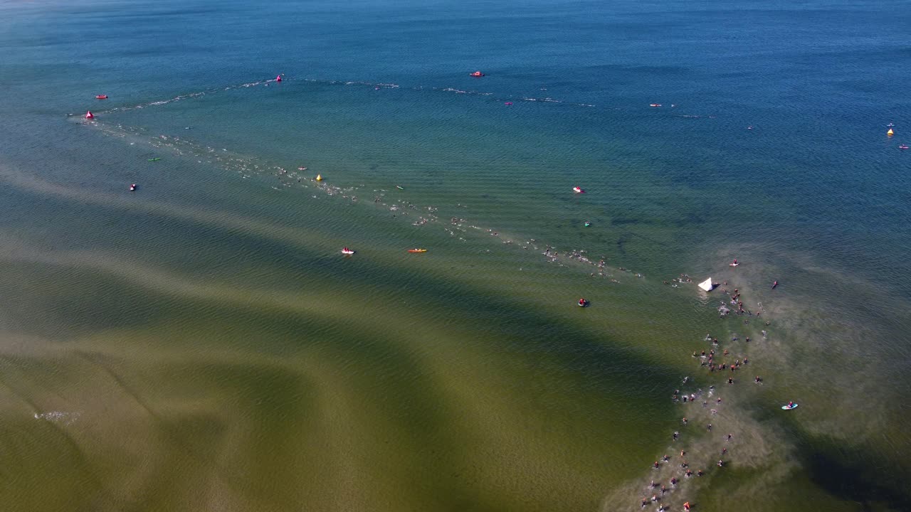 드론은 트라이애슬론 대회 기간 동안 탈린의 개방된 물에서 수영하는 선수들의 공중 사진을 찍고, 근처에 지원 보트가 있습니다.