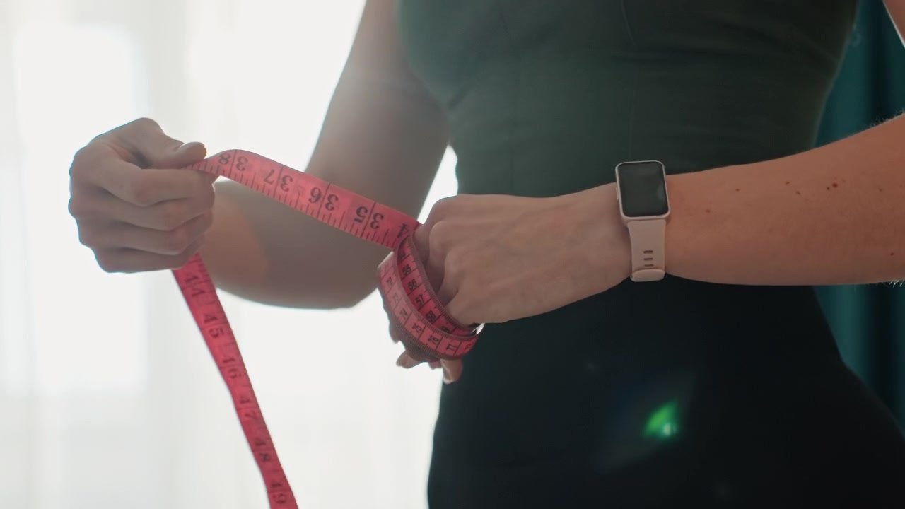 Closeup of fitness progress. Measuring waist with pink tape following workout routine. Individual monitors waist measurements using tape and smartwatch after physical activity session