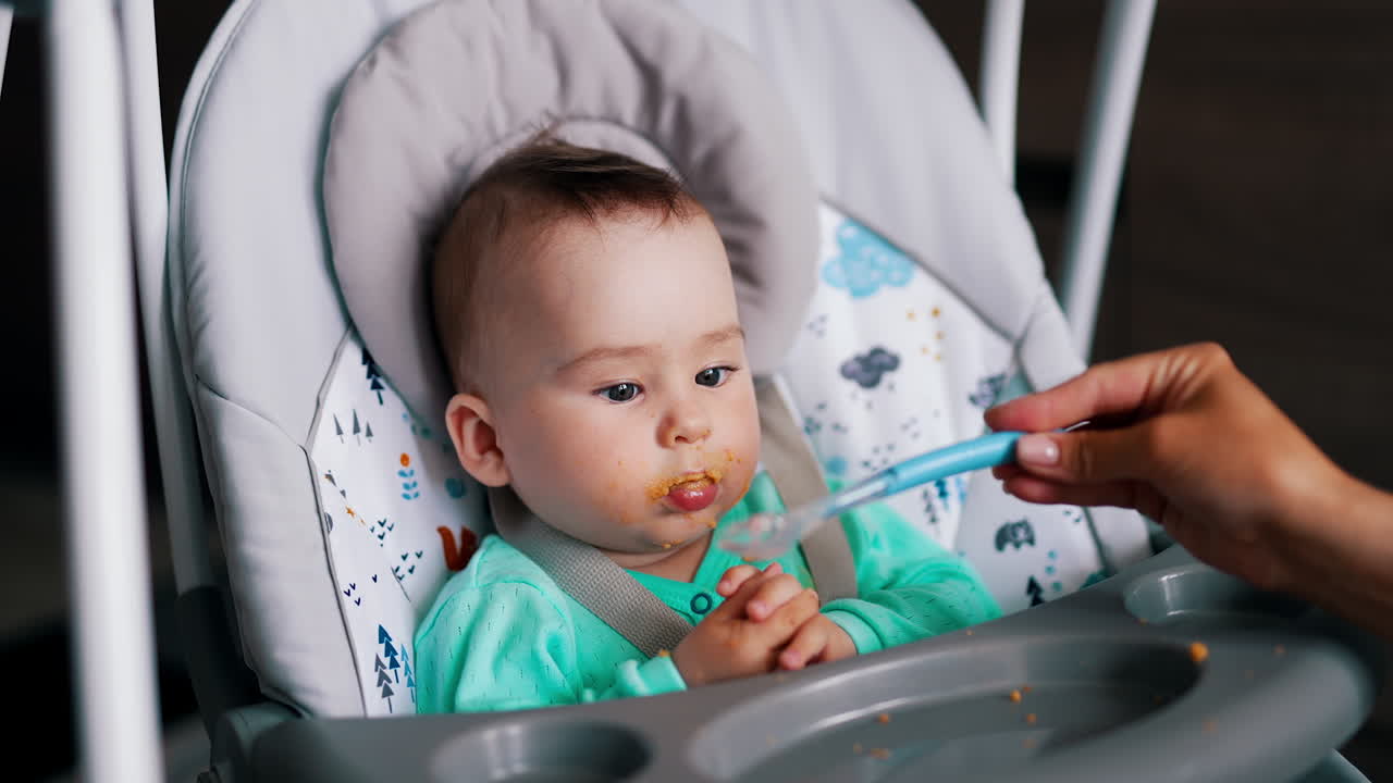 Lovely kid with dirty mouth sits in a baby chair. Mother's hand is giving a spoon to her son. Close up.