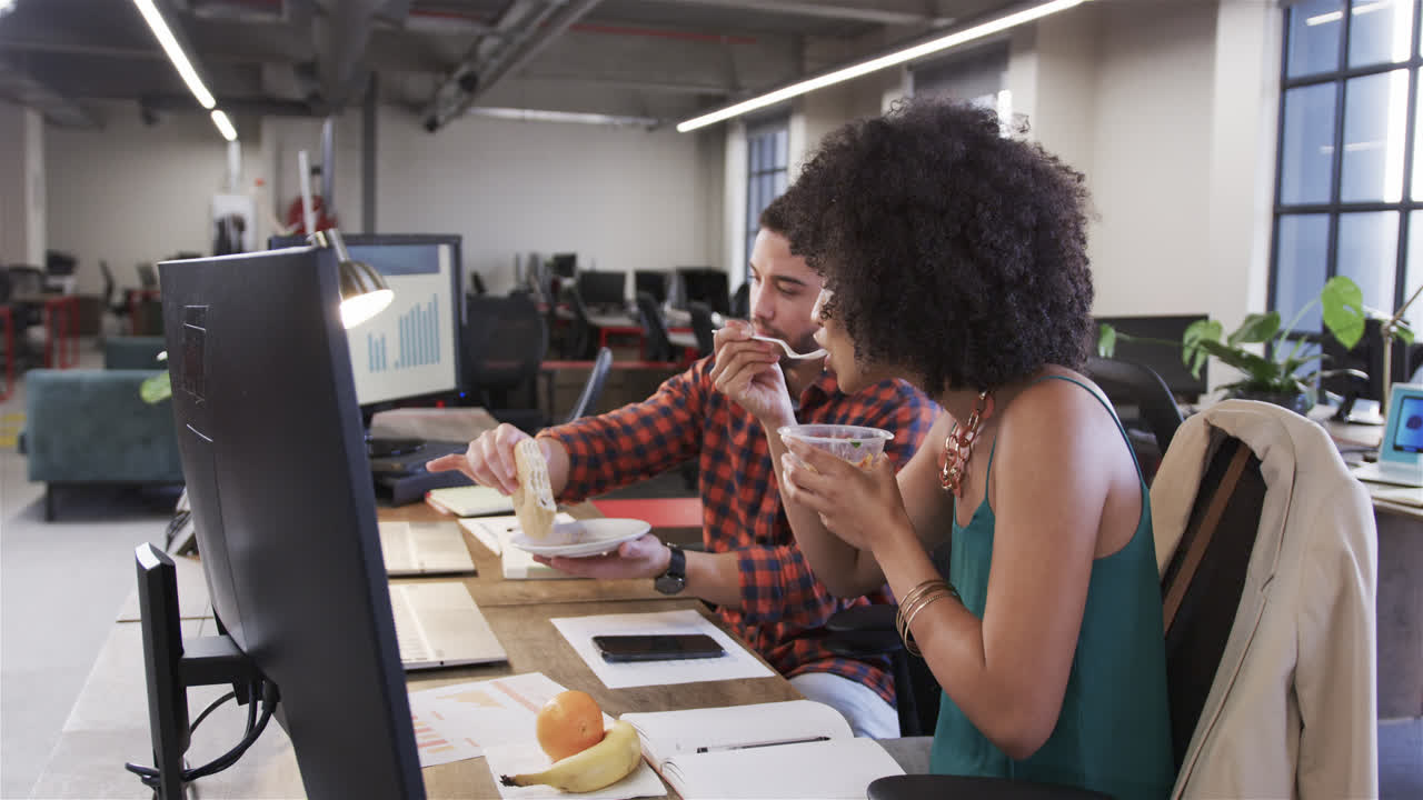 Happy diverse male and female colleagues sitting at desk eating and talking in office, slow motion