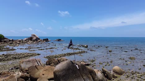 A pirate flag waves on a rocky shoreline at Lamai Beach, Koh Samui, Thailand. Turquoise water, clear blue sky, and scenic coastal landscape