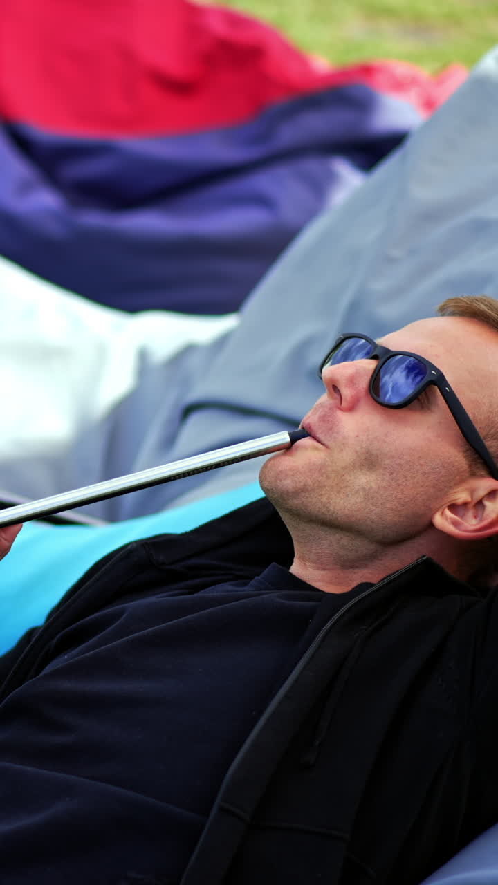 Relaxed man in black clothes and sunglasses lies in bean bag chair outdoors. Resting man smoking hookah producing smoke. Vertical video