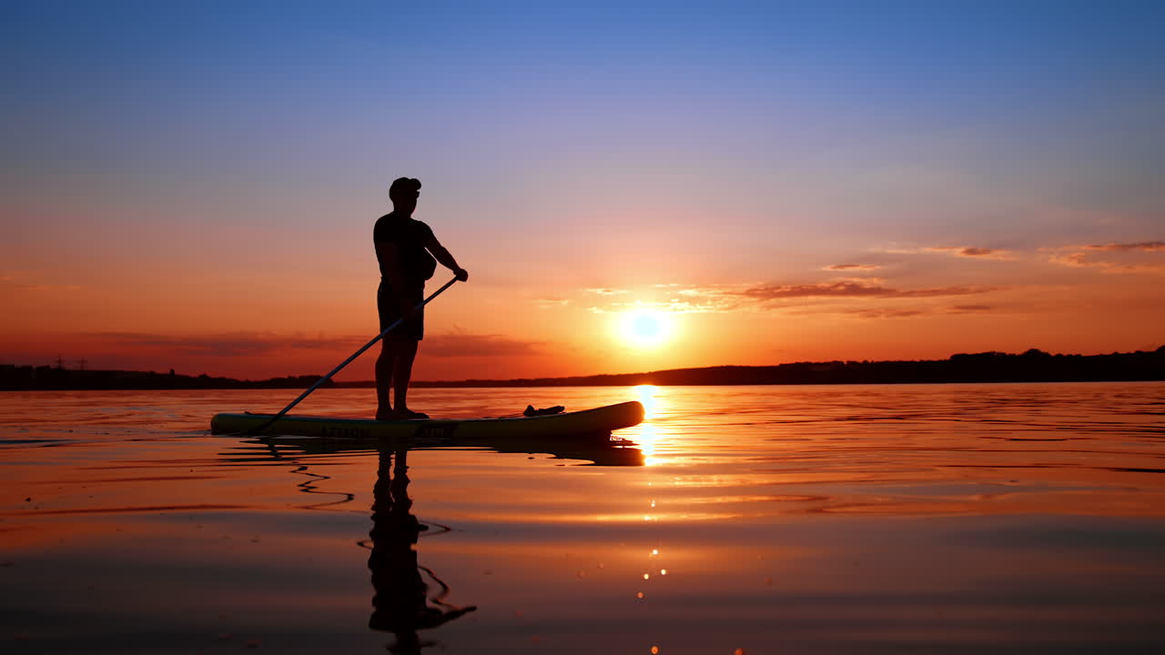 Tranquil evening on the river at sunset practicing sup boarding. Man in cap rows with an oar moving by the water on the sup board.
