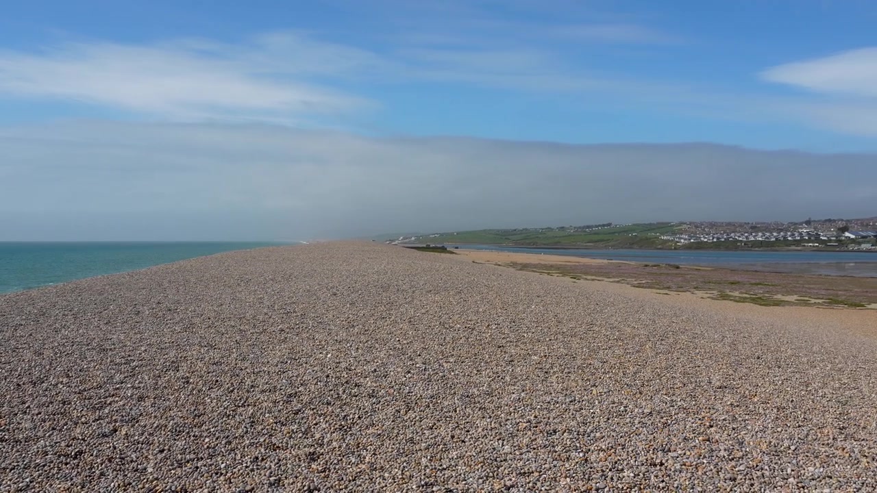 Una ripresa aerea cinematografica che mostra la vasta spiaggia di ciottoli di Chesil Beach a Weymouth, Dorset. Cattura la geografia costiera unica e il mare calmo sotto un cielo blu