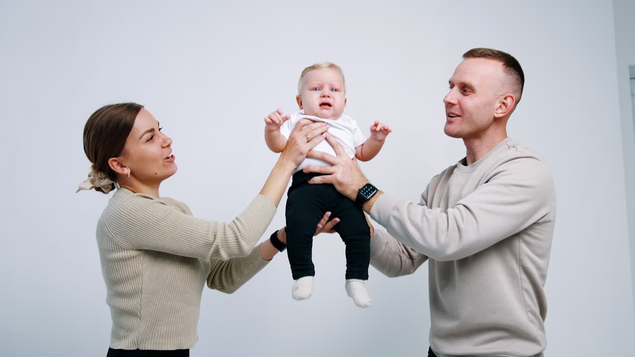 Caucasian parent are tossing their crying son. Married couple trying to calm sown their baby White backdrop.