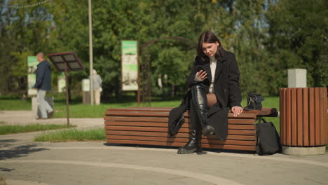 mujer sentada en un banco del parque con abrigo y botas negras, desplazando el teléfono inteligente, relajándose al aire libre, luz solar suave, fondo borroso con persona caminando y vegetación, bolso a su lado