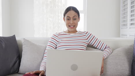 happy biracial woman sitting on sofa using laptop for video call, slow motion