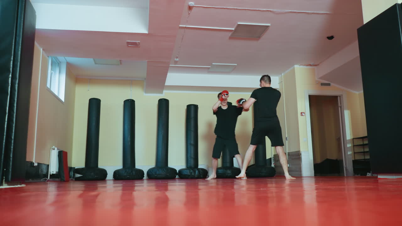 Atletas entrenando en un gimnasio de artes marciales; uno en posición defensiva con guantes rojos, agachado, mientras que el oponente está de frente, listo para atacar. Ambos descalzos sobre una colchoneta roja de entrenamiento, rodeados de sacos de boxeo.