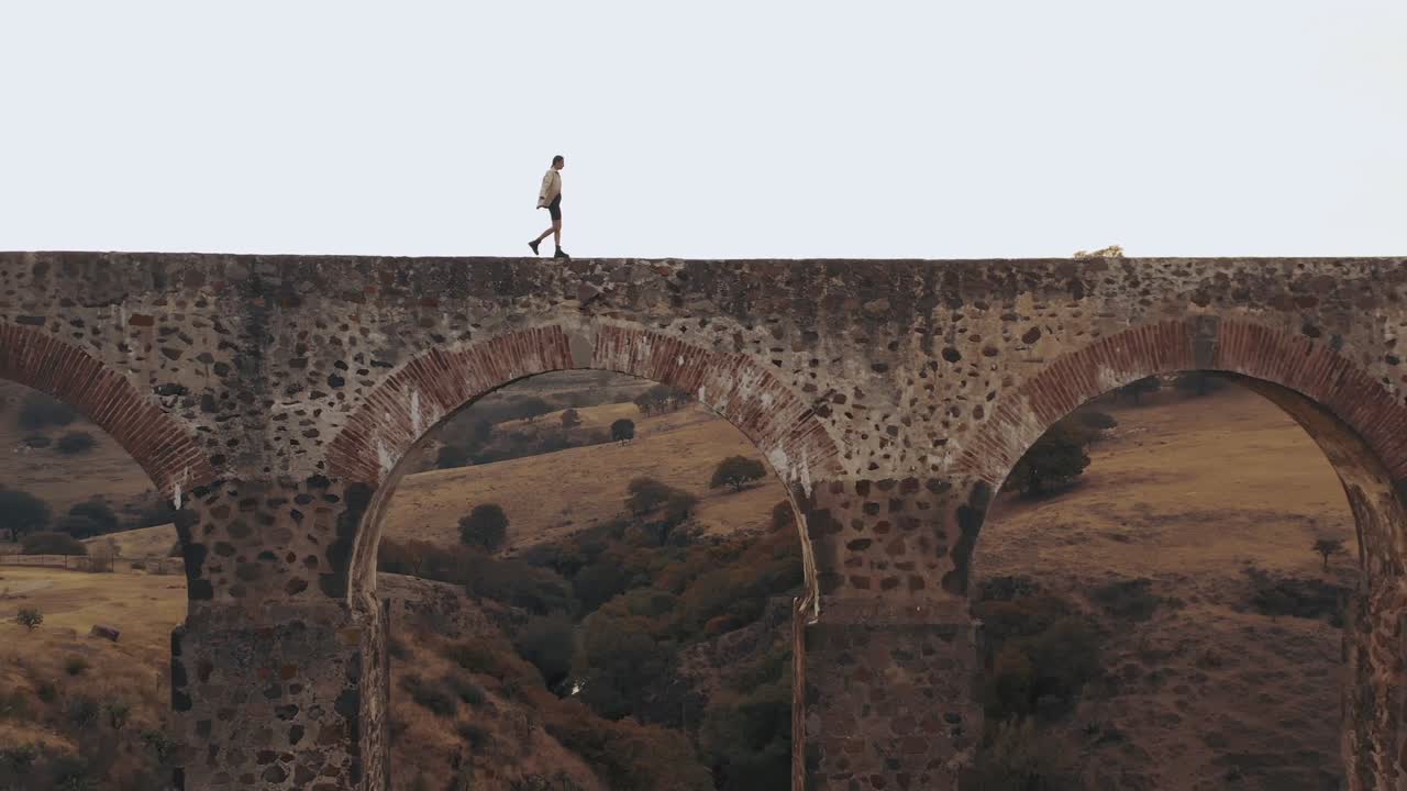 ariel fotografió a un hombre elegante y cariñoso caminando por un pequeño puente en italia en un caluroso día de verano por la tarde.