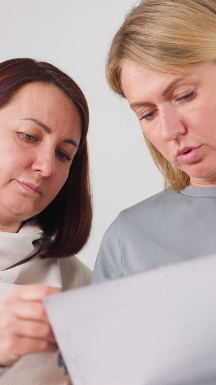 Due donne che esaminano un foglio durante una discussione collaborativa; partner creative che confrontano appunti su una stampa perforata, gesticolano e si concentrano, sfondo neutro da studio, espressioni coinvolte