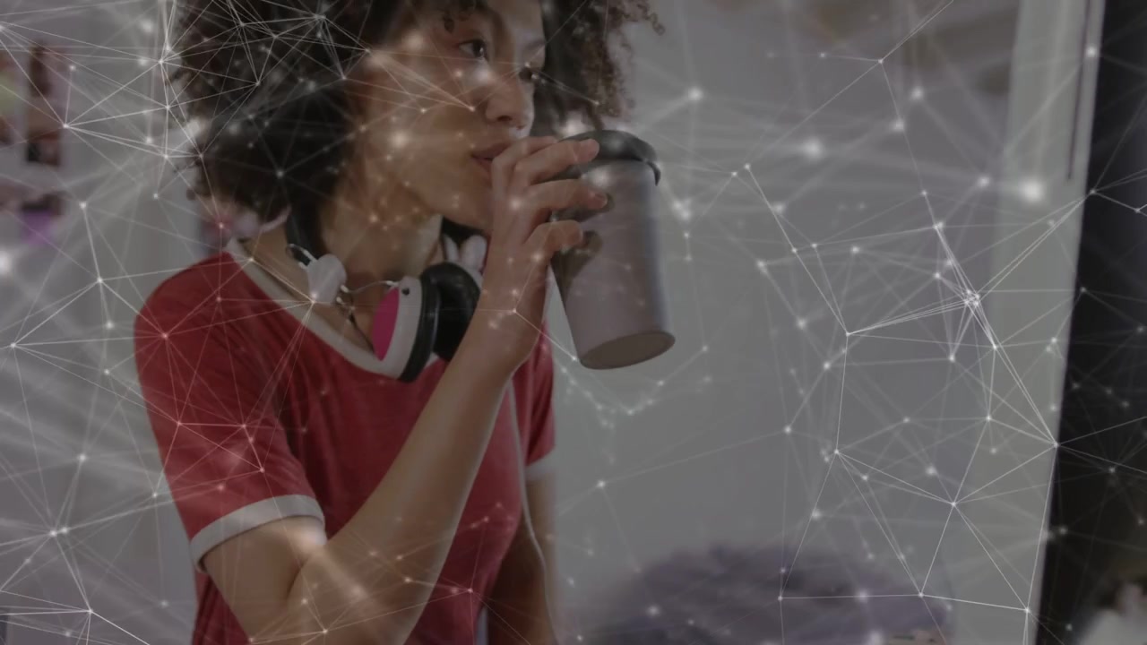 Woman reaching for tumbler, lifting, sipping while working on monitor with network overlay in tech