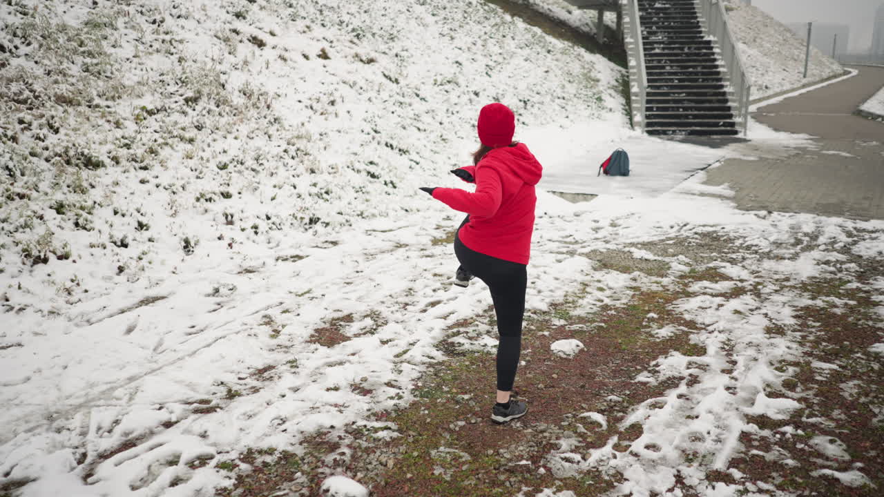 코치는 야외에서 활기찬 겨울 운동을 하며, 계단 근처의 눈 인 땅에서 다리를 들어 올리고, 빨간 후디와 검은 장갑을 입고, 눈 은 언덕, 경로, 안개가 가득한 분위기로 둘러싸여 있습니다.