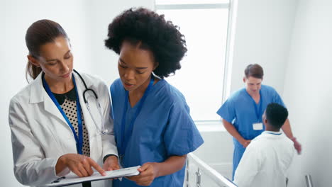 Female-Doctor-And-Nurse-With-Clipboard-Discussing-Patient-Notes-On-Stairs-In-Hospital-Building