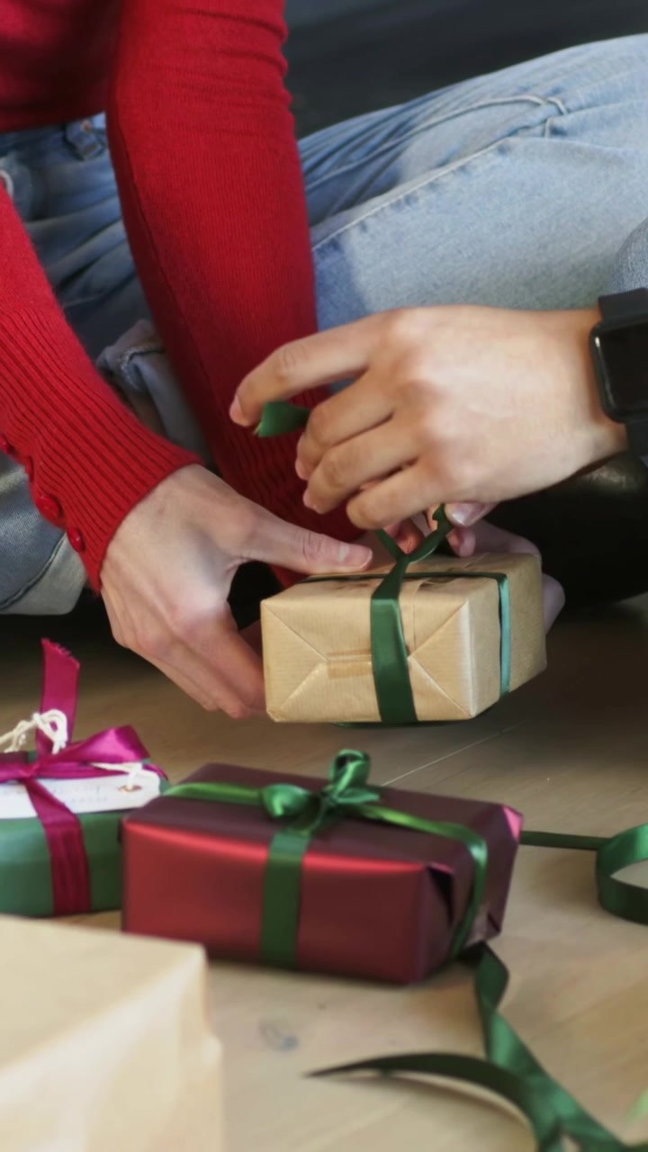 Video verticale: Mentre lega un maglione rosso, un aiutante si unisce tirando un nastro su una scatola di cartone kraft a casa per i regali.