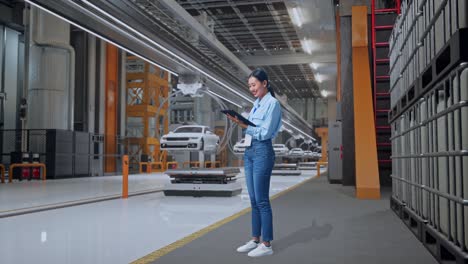 vista lateral completa del cuerpo de una trabajadora profesional asiática de pie con su tableta en una fábrica de automóviles ev, instalación industrial de alta tecnología, trabajando continuamente con su tablita