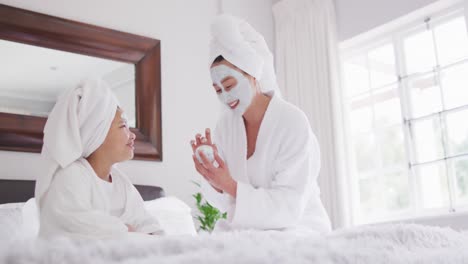 video of happy asian mother and daughter in robes moisturizing faces and having fun