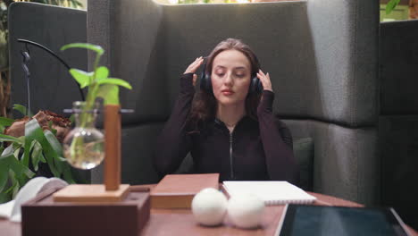 lady with blond hair tilts head back while putting on headset and prepares to open book on wooden table with indoor plant decoration, creating a cozy and focused atmosphere in modern setting