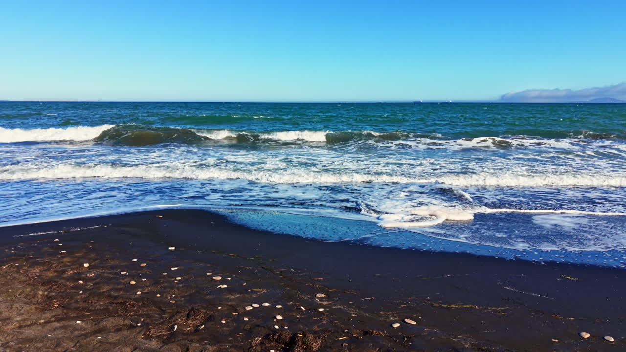 Scena di spiaggia con sabbia nera vulcanica e onde sotto un cielo sereno alla luce del pomeriggio.