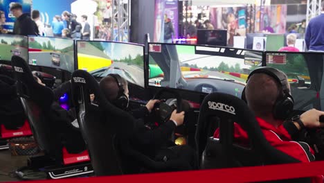 row of men sitting in front of screens sitting in a driver seat with steering wheel and wearing headsets playing a racing game tournament on a convention event at lgx luxembourg