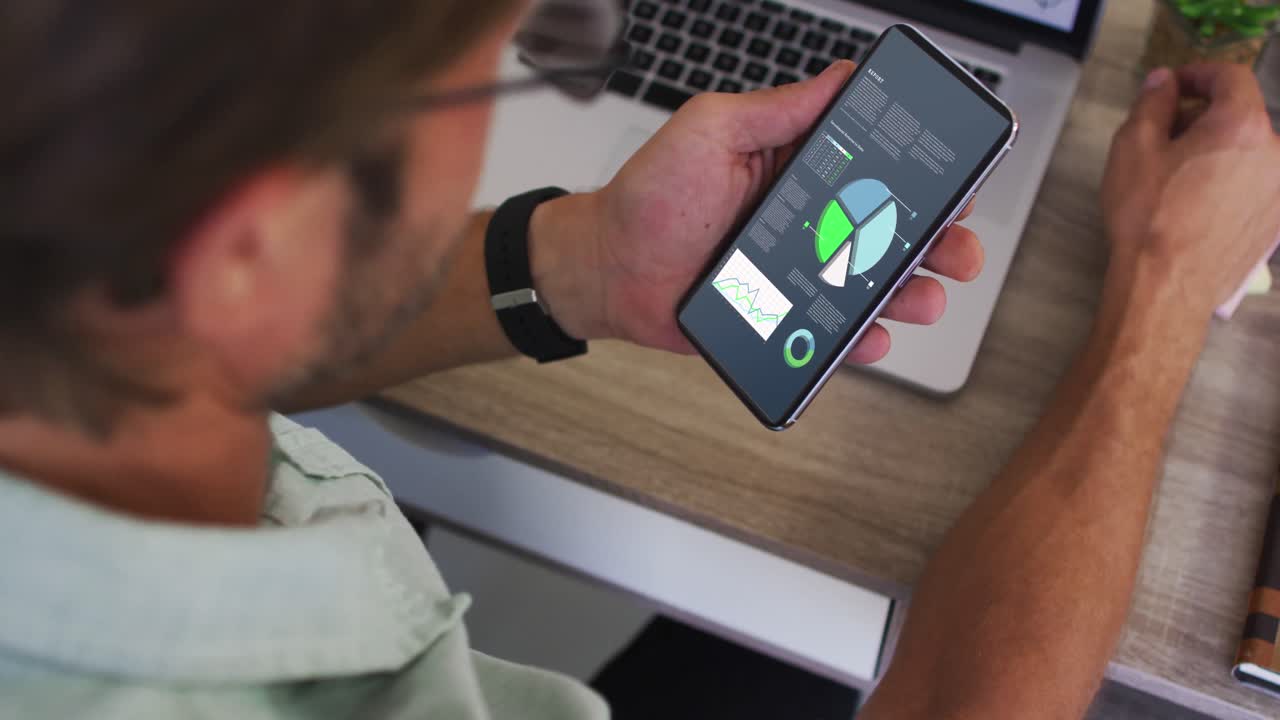 hombre caucásico sentado en un escritorio usando un teléfono inteligente con estadísticas en la pantalla