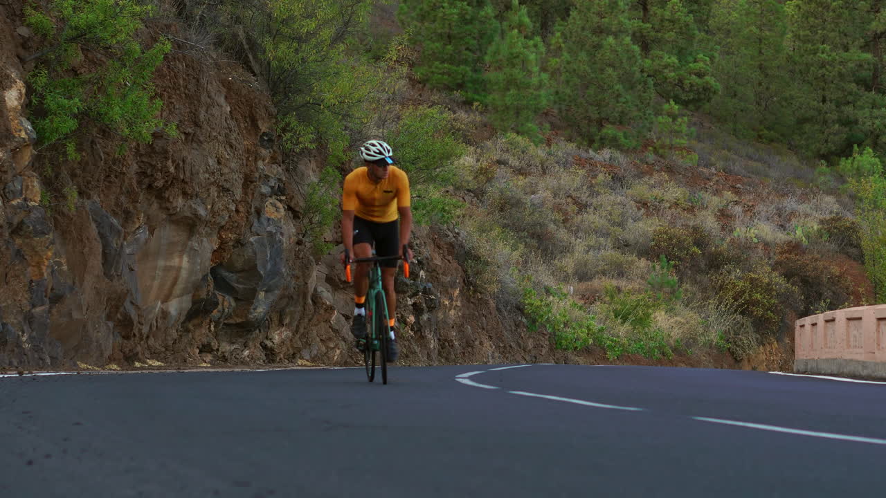 A man wearing a yellow t-shirt is cycling on a road situated at a high altitude in the mountains using a sports road bike