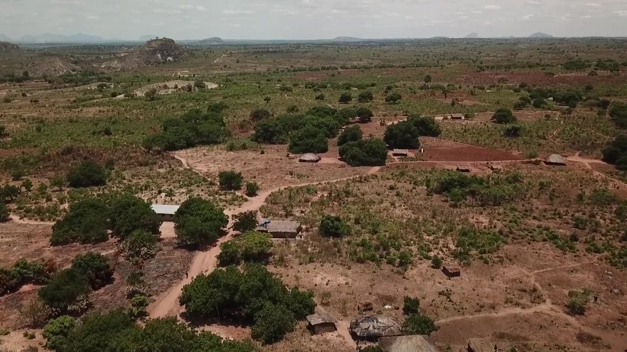 Lenta discesa aerea su un villaggio remoto nel Mozambico settentrionale, con case sparse, sentieri sterrati e un paesaggio di savana secca.