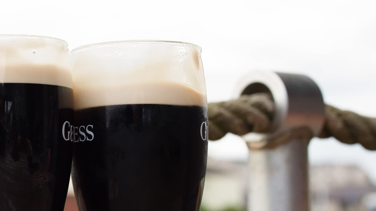 Two people clink dark beer glasses outdoors, natural daylight, shallow depth of field, steady camera