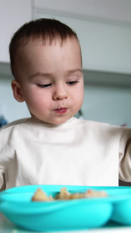 adorable caucasian baby in white sweater eating at home. kid sits at table with blue plate and mom gives a full spoon to her son. blurred backdrop. vertical video
