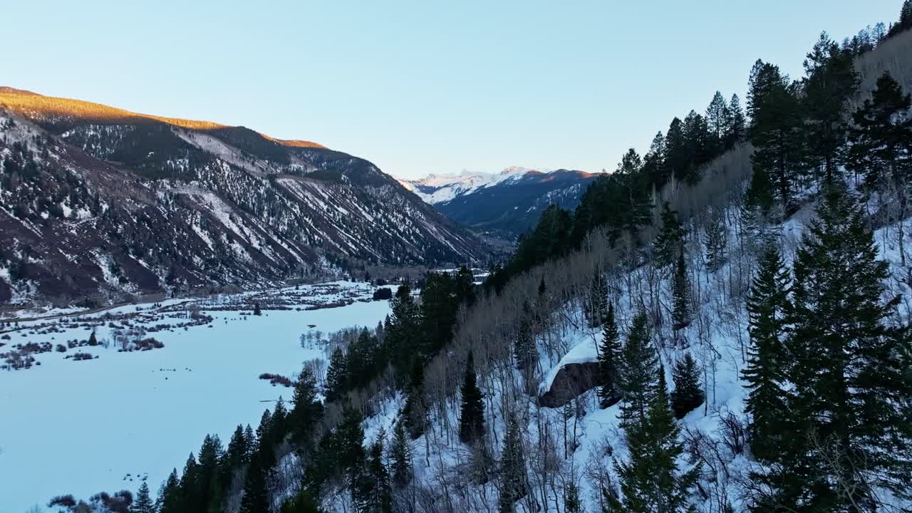 Mountain slopes curve gently into valley with forest shadows deepening during soft sunset hour, aerial dolly
