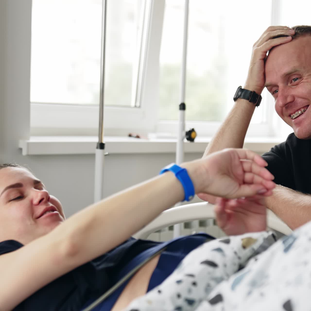 Um homem está sentado na cama de uma mulher. Marido e esposa grávida conversam e riem enquanto a mulher é examinada com um dispositivo médico.