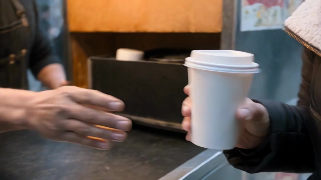 Mano del venditore che porge una tazza bianca a un carretto ambulante, servendo un cliente in cappotto, pendolari che attraversano.