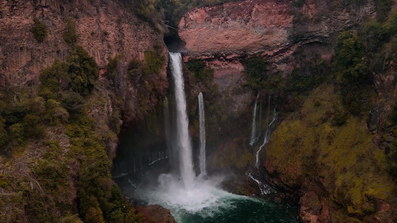 Backward drone aerial of waterfall with mist into turquoise pool, Siete Tazas National Park, Ñuble, Chile with red rock cliffs and forest