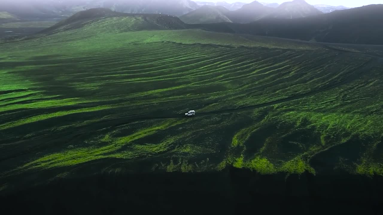 Aerial drone footage of a white 4x4 truck jeep driving on a rural and desolate nature road in the mountains at Greenland or Iceland during a cloudy day. Mossy and green fields with sharp edges around.