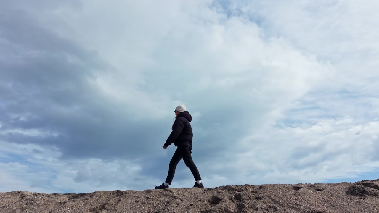 Young woman walking on a sandy hill on the seashore. Woman in a jacket walking on the street in autumn. Woman having fun and walking on the sand.