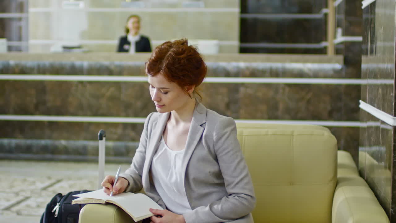 Tracking Of Young Businesswoman Sitting In Armchair And Writing In Organizer While Businessman Walking Through Hotel Lobby In Slow Motion