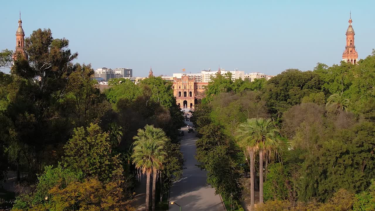 Smooth aerial dolly‑in moves forward along a straight park avenue through Parque de Maria Luisa, slowly revealing the central building and twin towers of Plaza de España in Seville, Andalusia, Spain