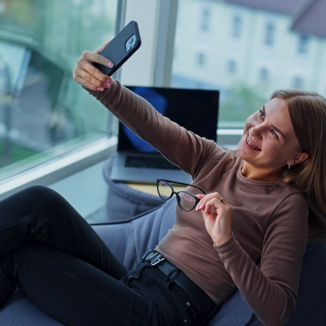 Positive smiling young woman half-lying in a bean bag chair with phone in stretched hand. Lady having video chat during break from work. Blurred backdrop