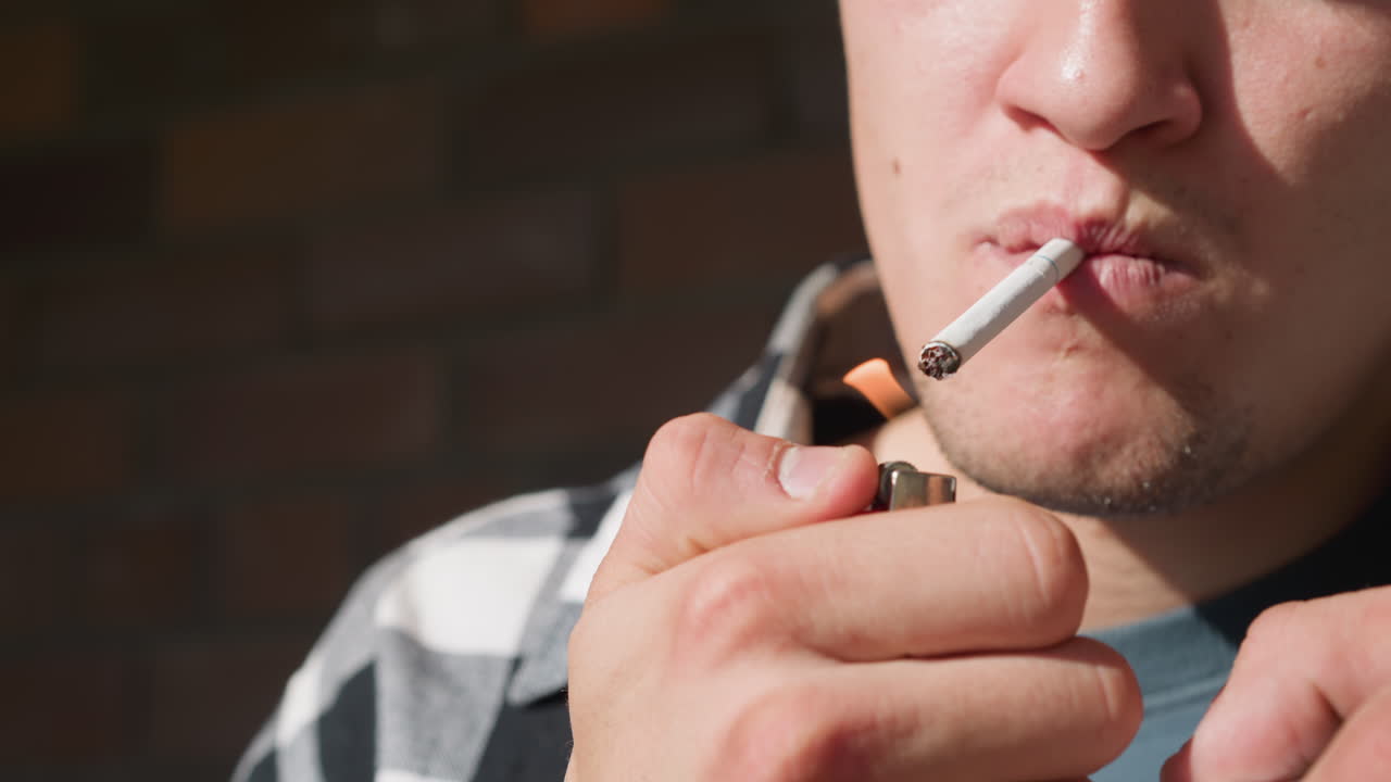 Close up of young man holding lighter and pack while lighting cigarette between lips, fingers flicking flame under tip, soft blurred brick wall and iron railing background
