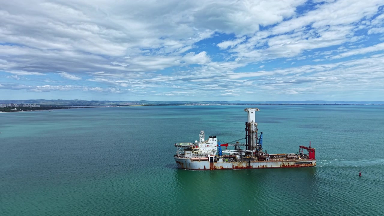 Industrial dredging vessel operates in open water near coastal area with clear skies and distant land in view