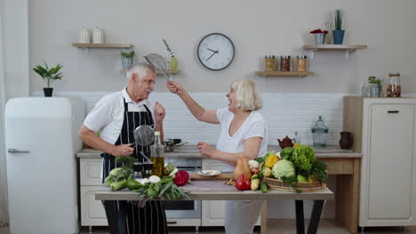 senior woman and man making a funny dance with strainers. dancing while cooking together in kitchen