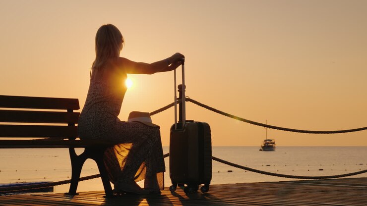 Une femme adulte, vêtue d'un sarafan d'été, est assise sur un banc de la plage et tient une valise à roulettes.