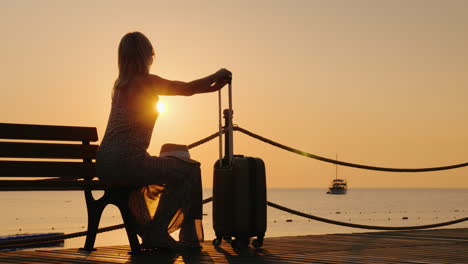 An-Adult-Woman-Sits-In-A-Summer-Sarafan-On-The-Beach-Sits-On-A-Bench-And-Holds-Onto-A-Suitcase-On-Wh