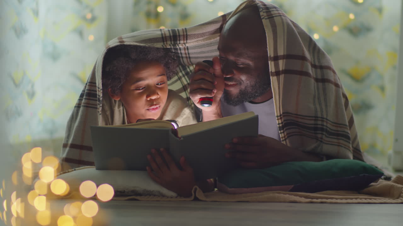 African American Dad and Son Reading Book on Bed